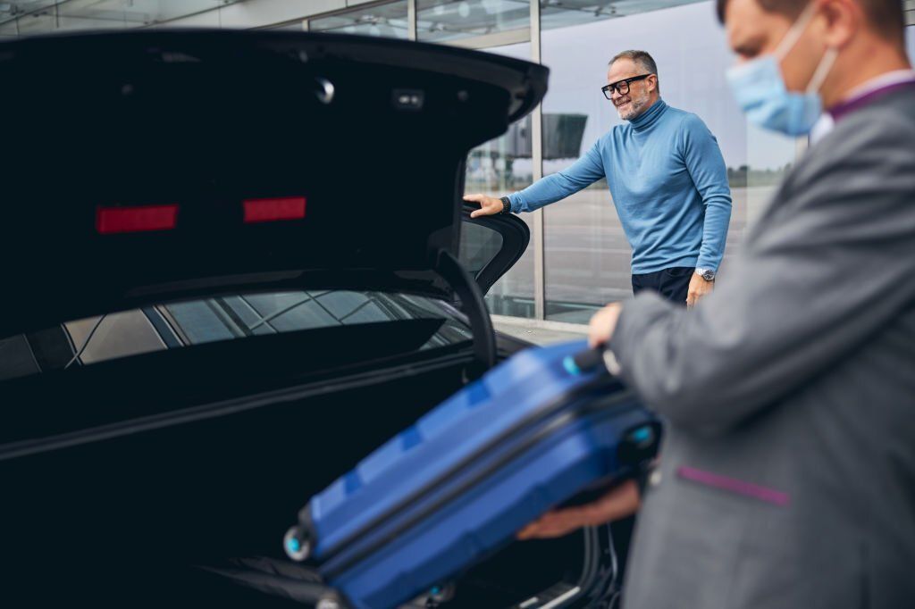 A smiling man wearing a blue shirt is about to ride a car as the chauffeur puts his small luggage in the trunk.