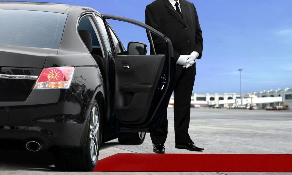 A man with white gloves stands beside a limo with open door by the red carpet.