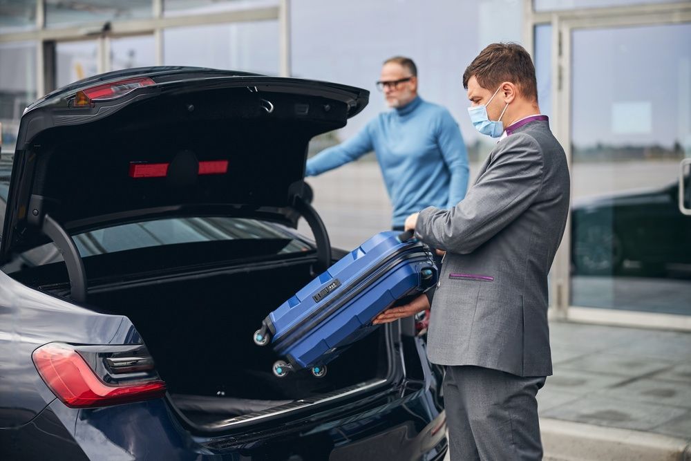 A uniformed chauffeur wearing face mask puts a small blue luggage of a male passenger in the car's compartment.