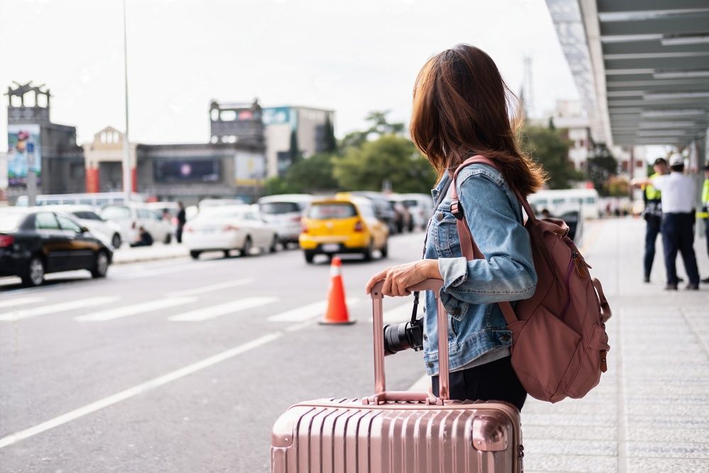 A woman holding a pink luggage stands outside the airport waiting for a ride.