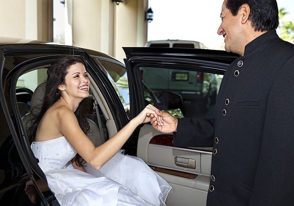 A smiling chauffeur takes the hand of a young and beautiful bride and helps her step out of the limo.