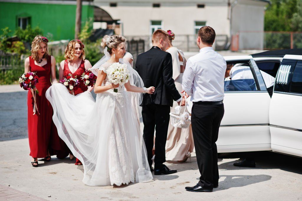 Two bridesmaids in red holds the train of a beautiful bride as the rest of the wedding entourage steps out of the limo.