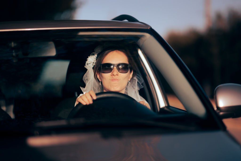 A frustrated-looking bride wearing sunglasses is driving a car.