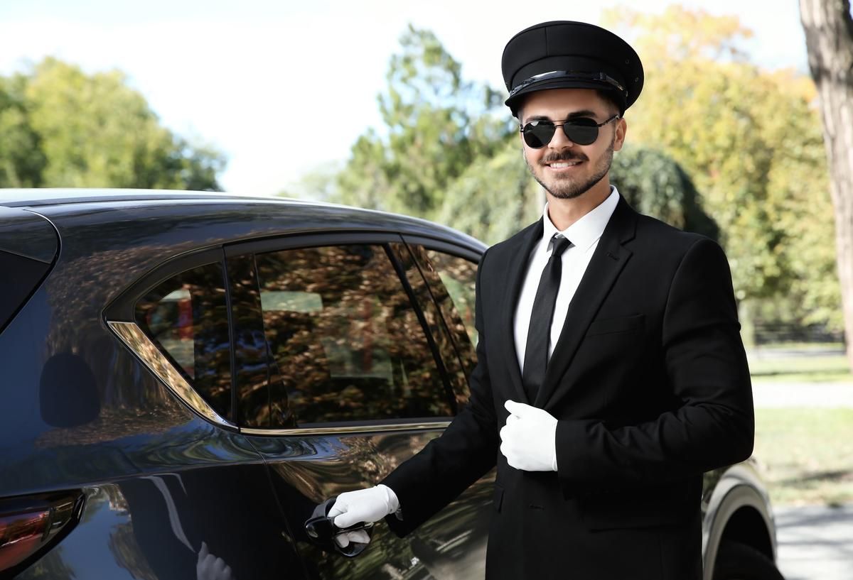 A well-groomed chauffeur smiles as he holds the door of a limo.