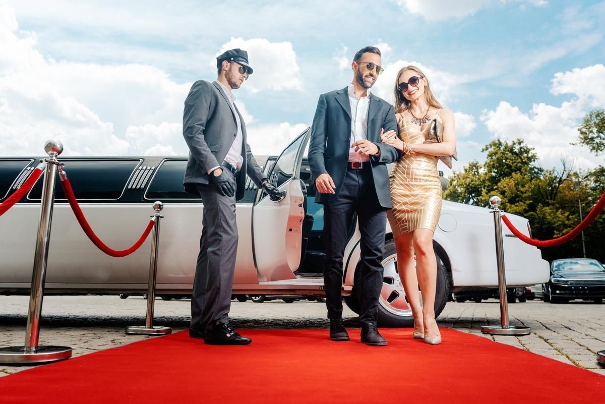 An elegant couple walks down the red carpet to an event while the chauffeur holds the door of a stretch limo.
