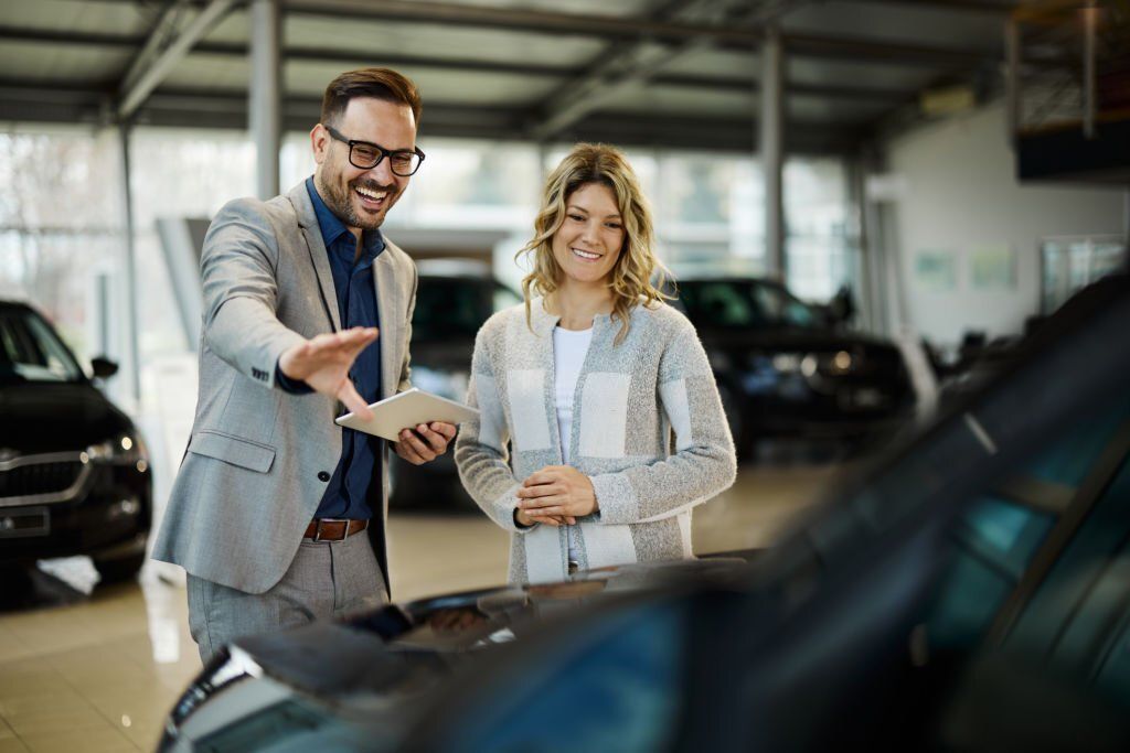 A happy car salesman showing a car to his customer in a showroom.