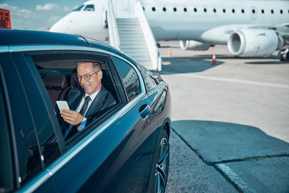 A businessman holding a mobile phone sits inside a limousine parked near a jumbo jet.