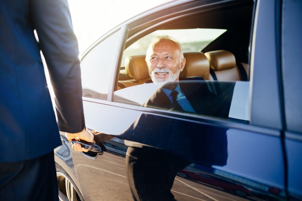 An elderly businessman is smiling at the chauffeur who is holding the door of the car for him.