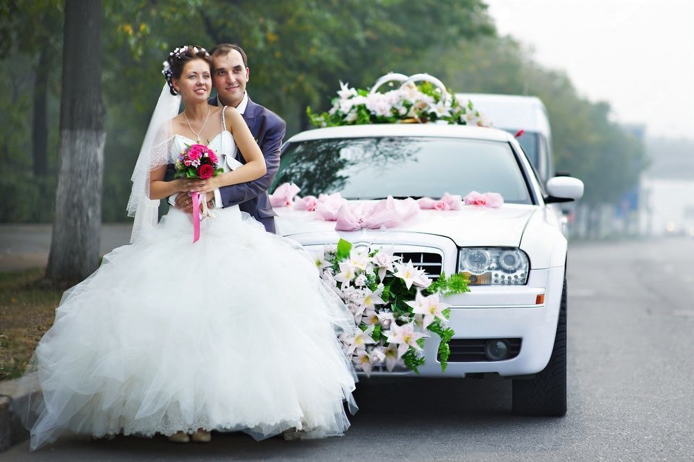 Newly married couple posing for a photo in front of a limousine decked with flowers.