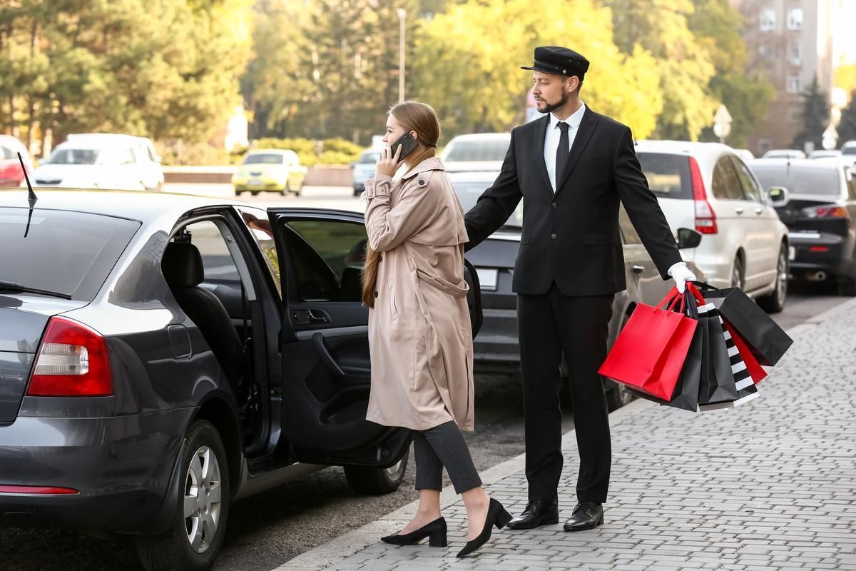 A woman is on the phone while a chauffeur holds the door of a car while carrying a number of shopping bags.