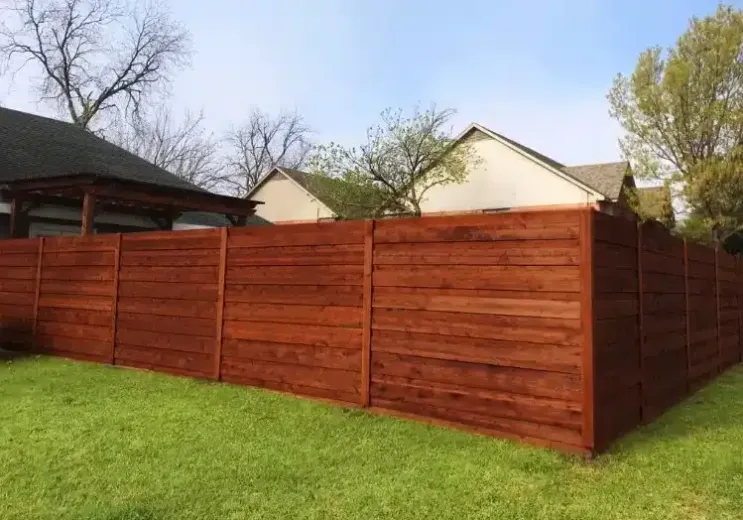 A stained wooden privacy fence in a backyard, with green grass and houses in the background on a sunny day.