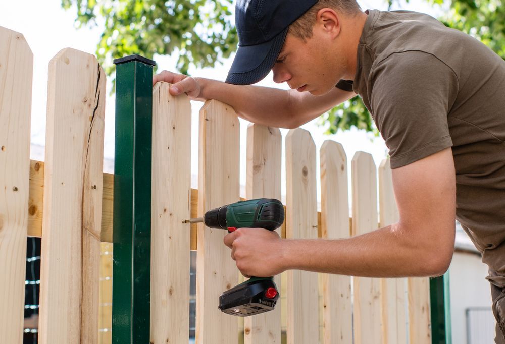 Man in a cap using a power drill to attach a wooden picket to a green fence post outdoors.