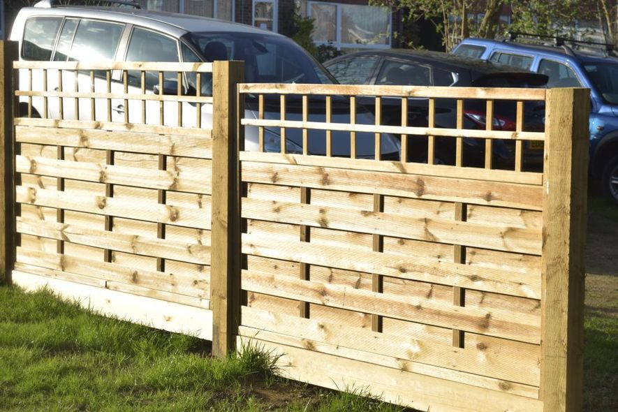 Wooden fence with woven plank design and lattice top section, in a grassy outdoor setting with parked cars in the background.
