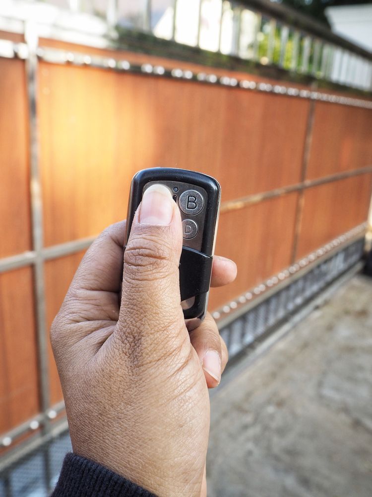 Person's hand holding a black remote control, pressing a button to open a brown gate.