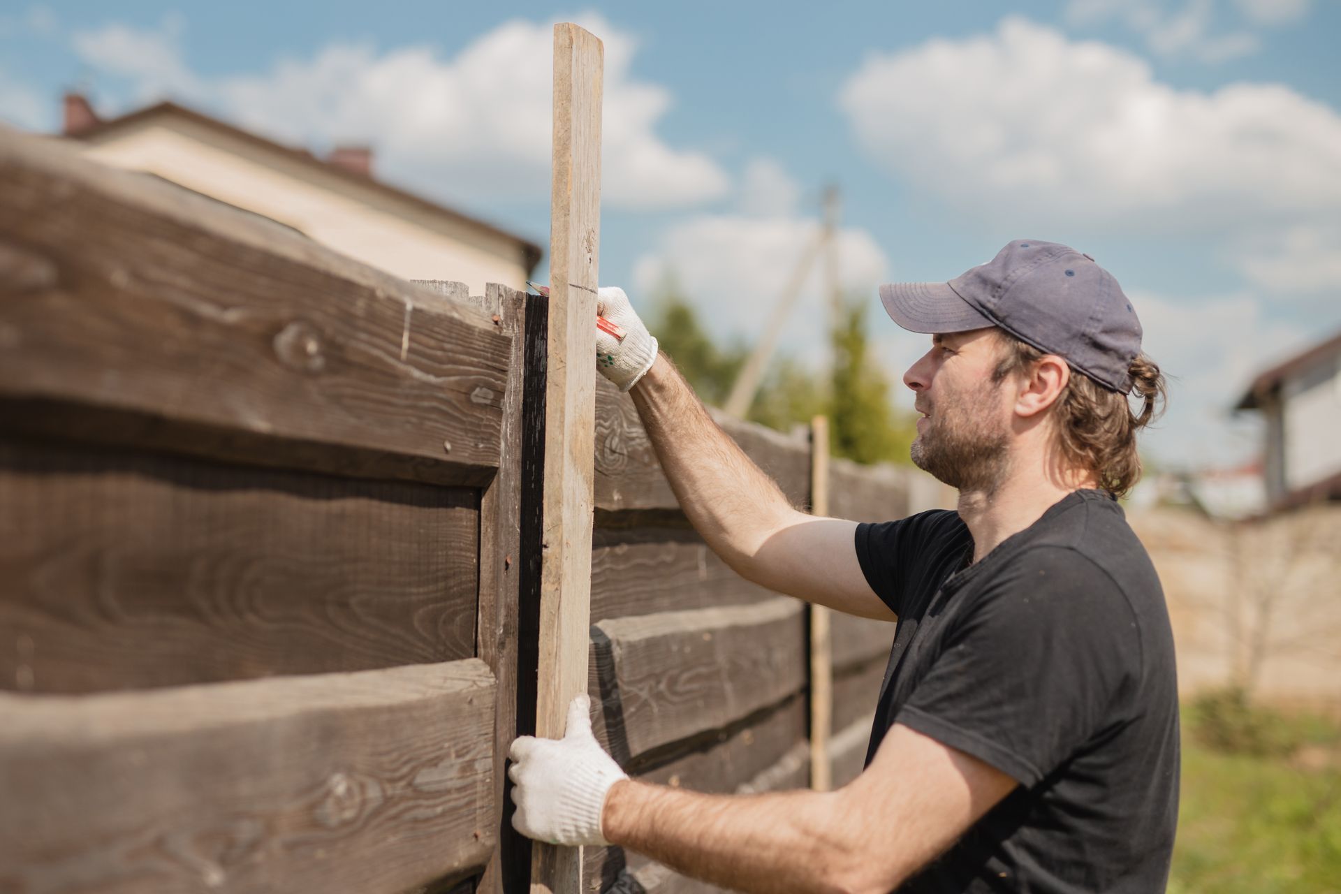 Man in cap and gloves installing wooden fence post outdoors.