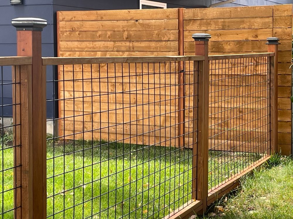 Person trimming a wooden fence with shears outdoors on a sunny day.