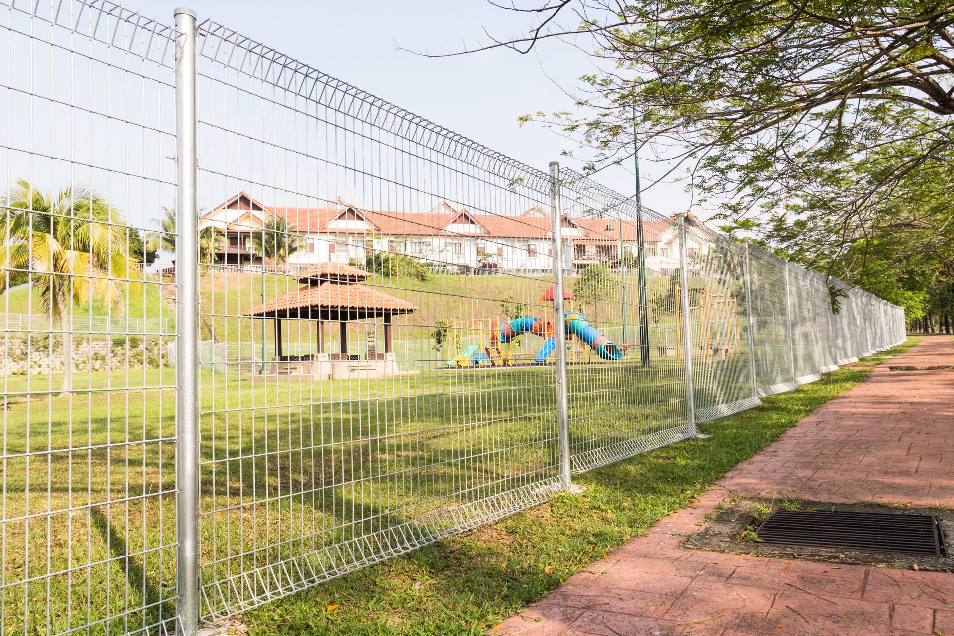 A chain-link fence borders a grassy area with a playground and houses in the background, alongside a red brick path.