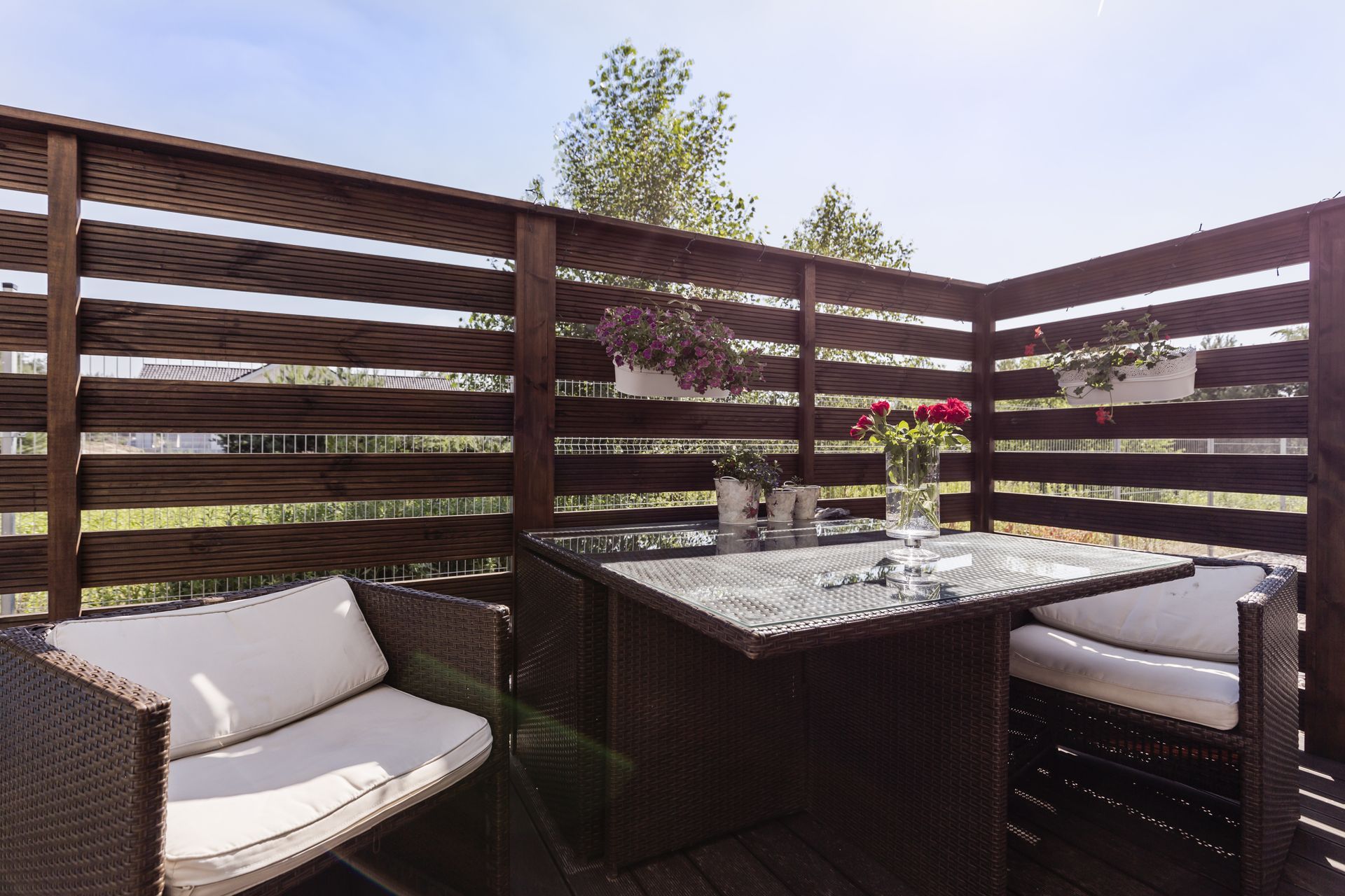 A patio with a dark brown wicker table and chairs, a wooden fence, and potted flowers, set against a sunny sky.