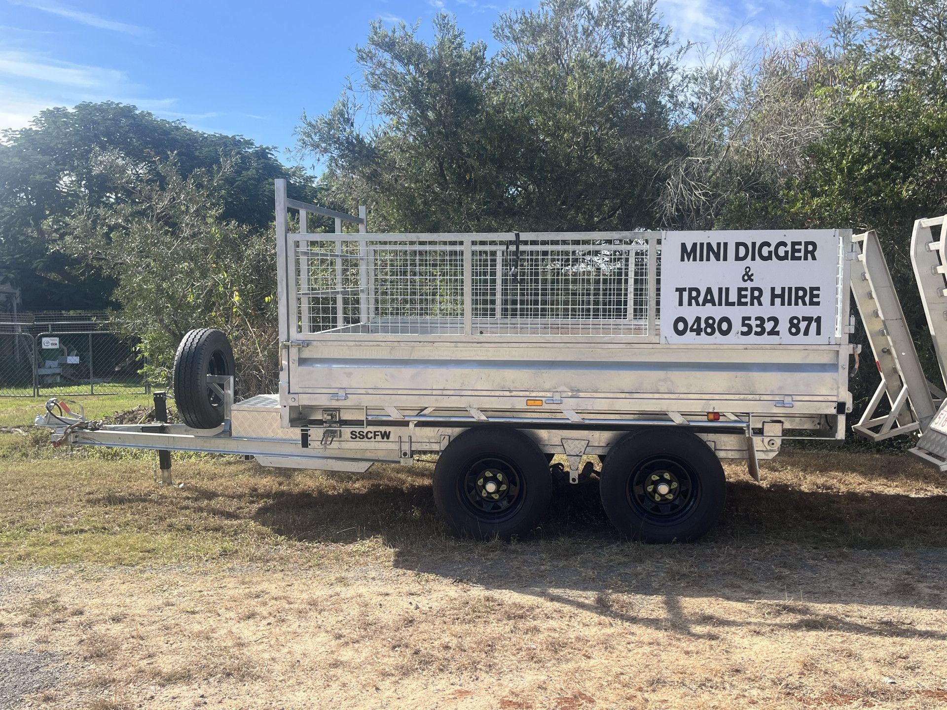 Trailer with NINI DIGGER & TRAILER HIRE sign; spare tire, two wheels, in outdoor setting.— Mini Digger and Trailer Hire in Atherton, QLD
