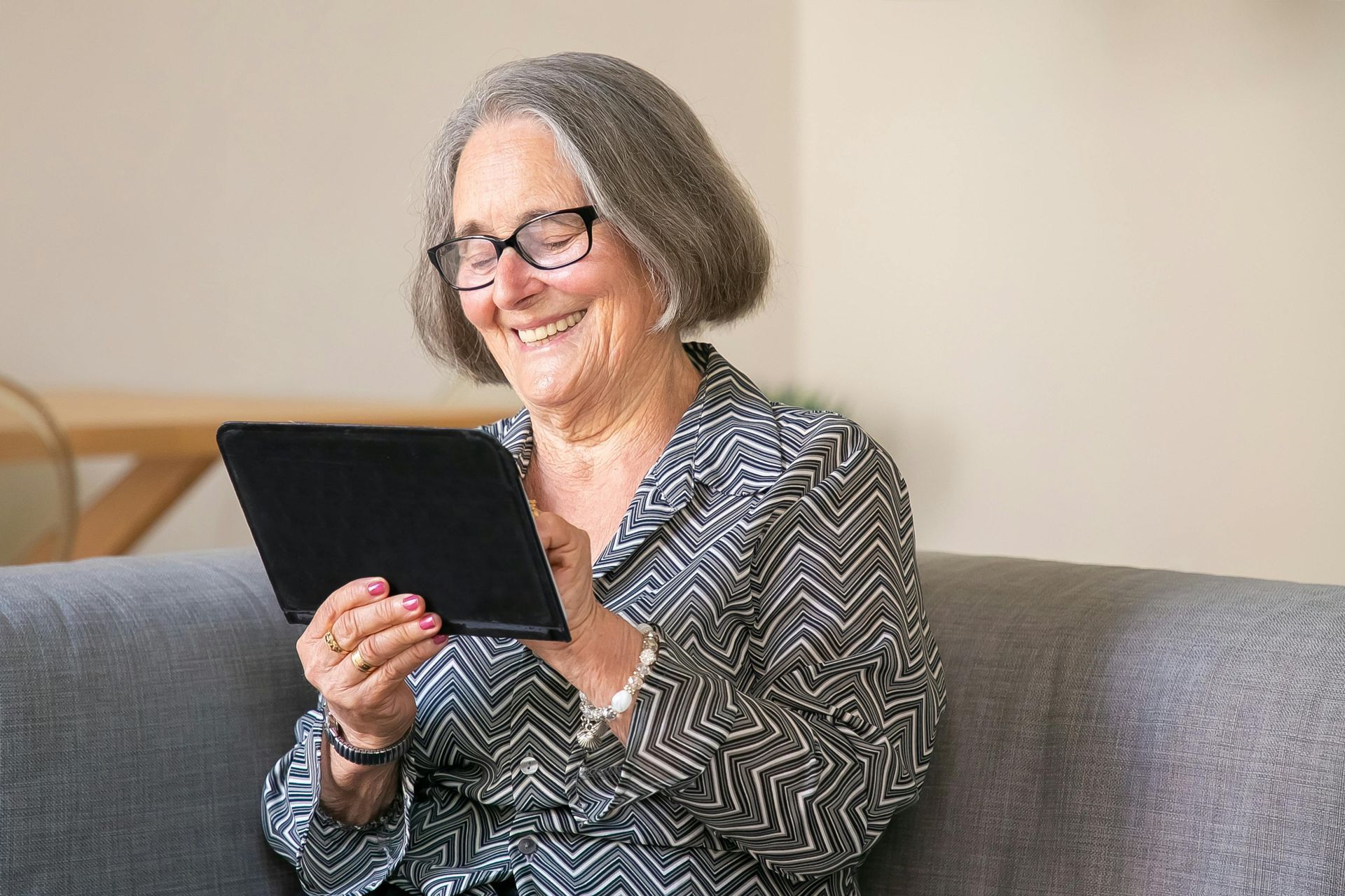 An elderly woman is sitting on a couch using a tablet computer.