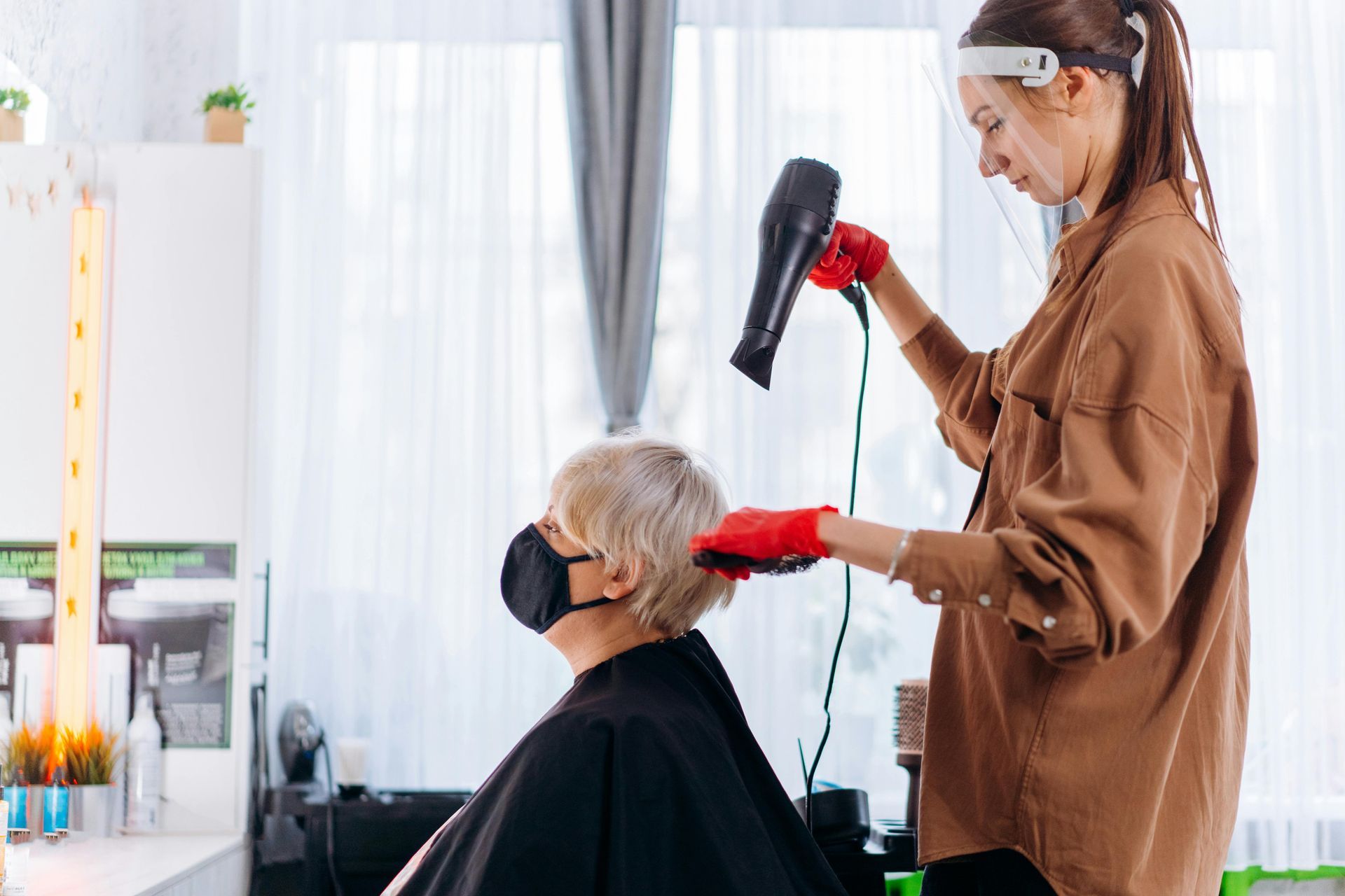 A woman wearing a mask is getting her hair blow dried by a hairdresser.