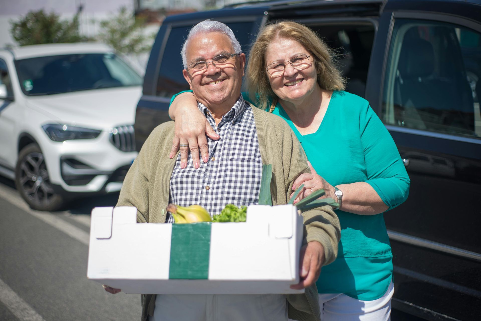 An elderly couple is standing next to a car holding a box of fruit and vegetables.
