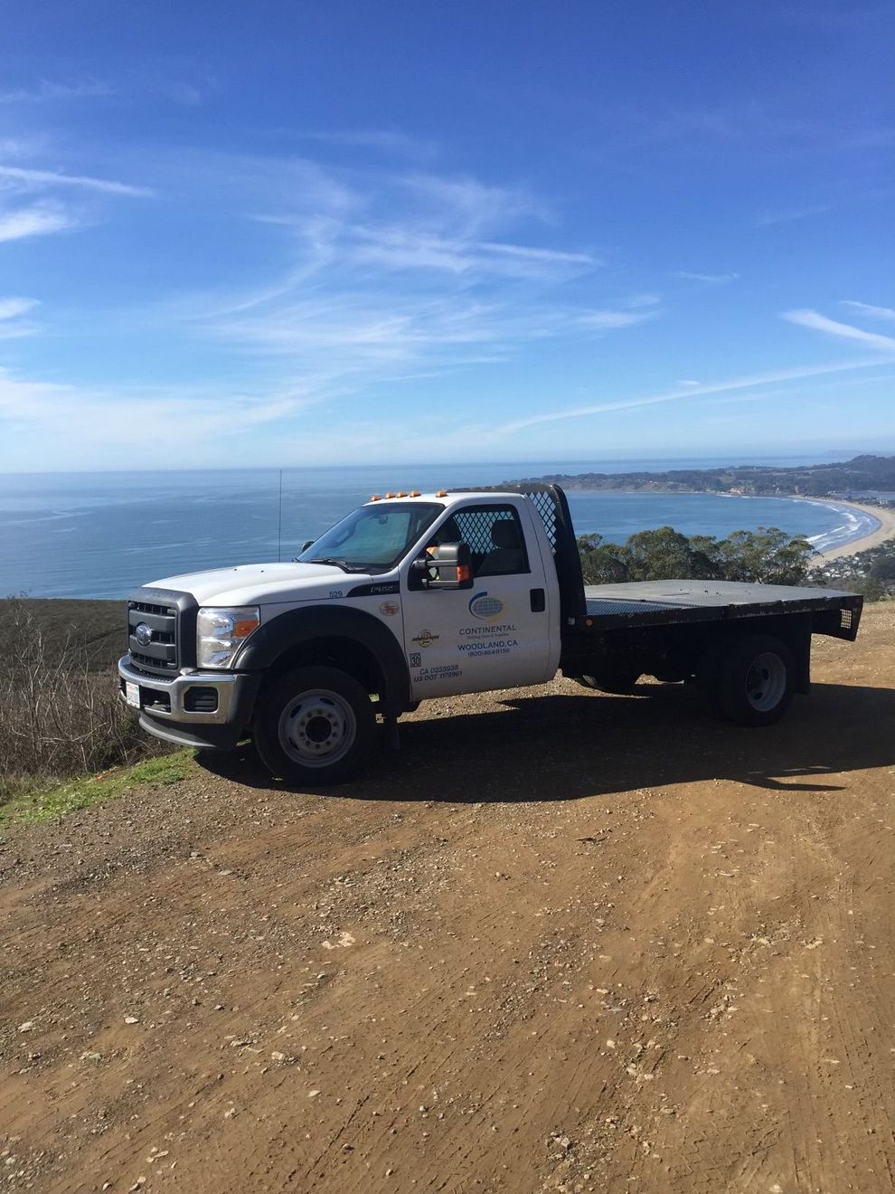 A white tow truck is parked on the side of a dirt road overlooking the ocean.