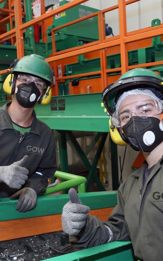Dos trabajadores con uniformes verdes, cascos y mascarillas levantan el pulgar en un entorno de fábrica.