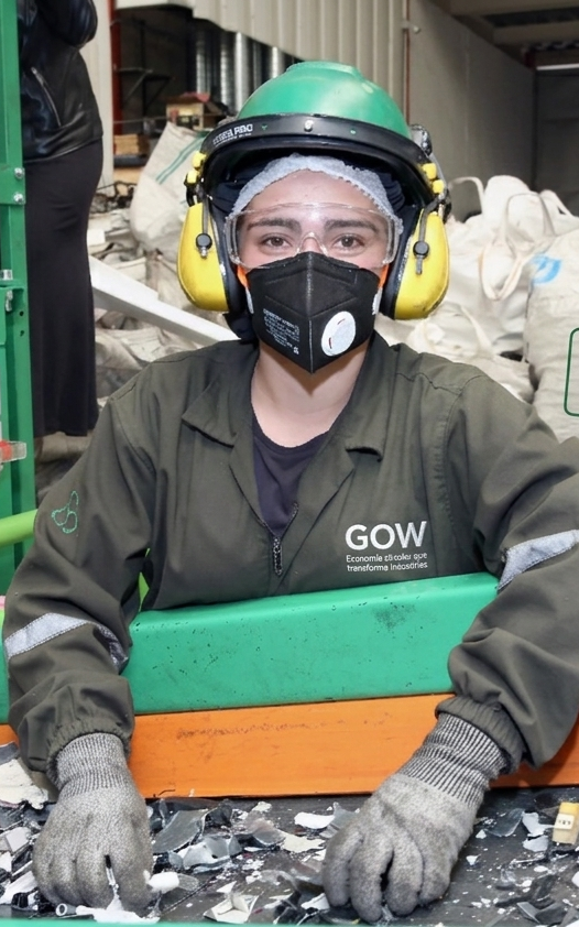 Obrero de fábrica con ropa verde, mascarilla, guantes y protección auditiva en una línea de producción manipulando materiales.