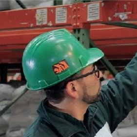 Un trabajador con casco verde y gafas se estira hacia arriba en un almacén industrial.
