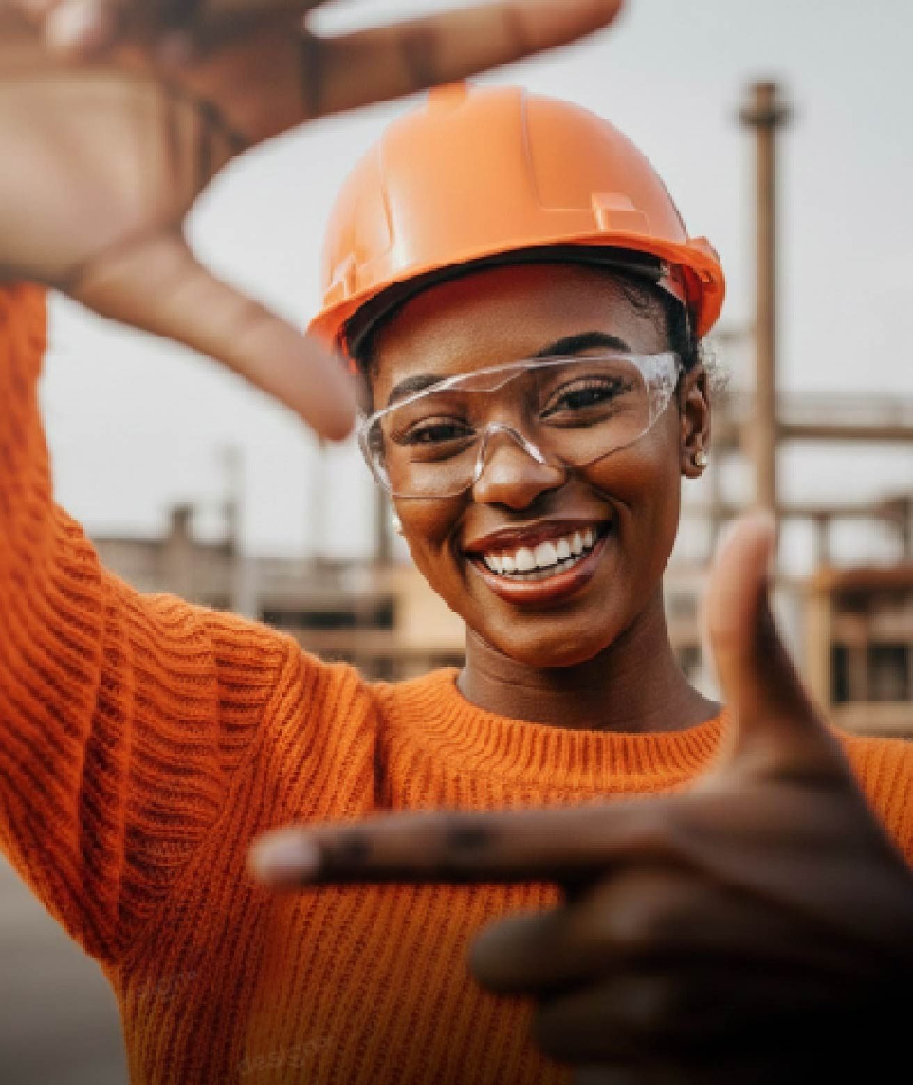 Un trabajador sonriente, con casco y suéter naranjas, en una obra industrial, fabrica un marco con sus manos.