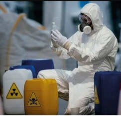 Una persona con traje de protección contra materiales peligrosos y respirador inspecciona un vial junto a barriles de residuos peligrosos.