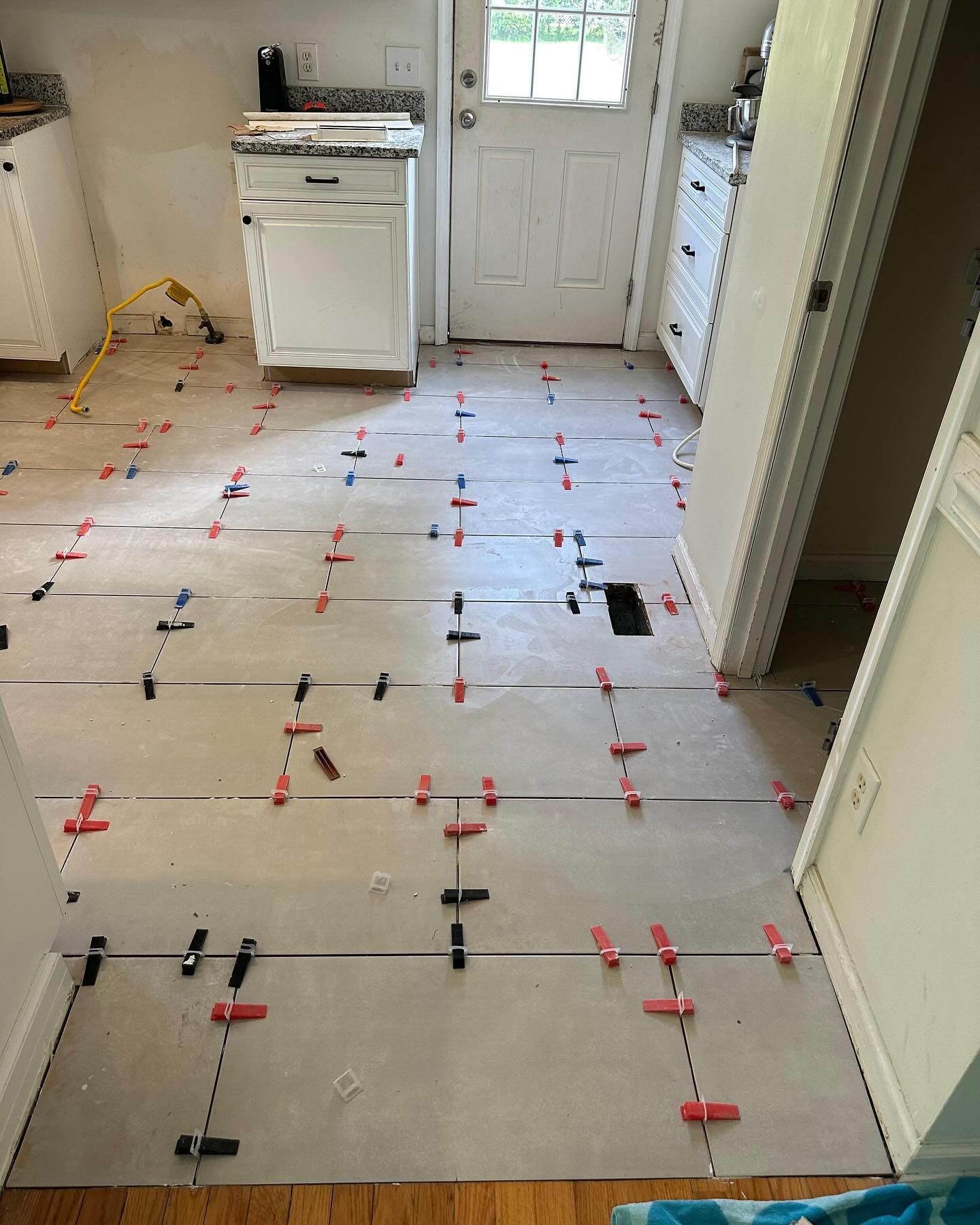 Kitchen floor being tiled, with gray tiles and red leveling clips leading to a white door.