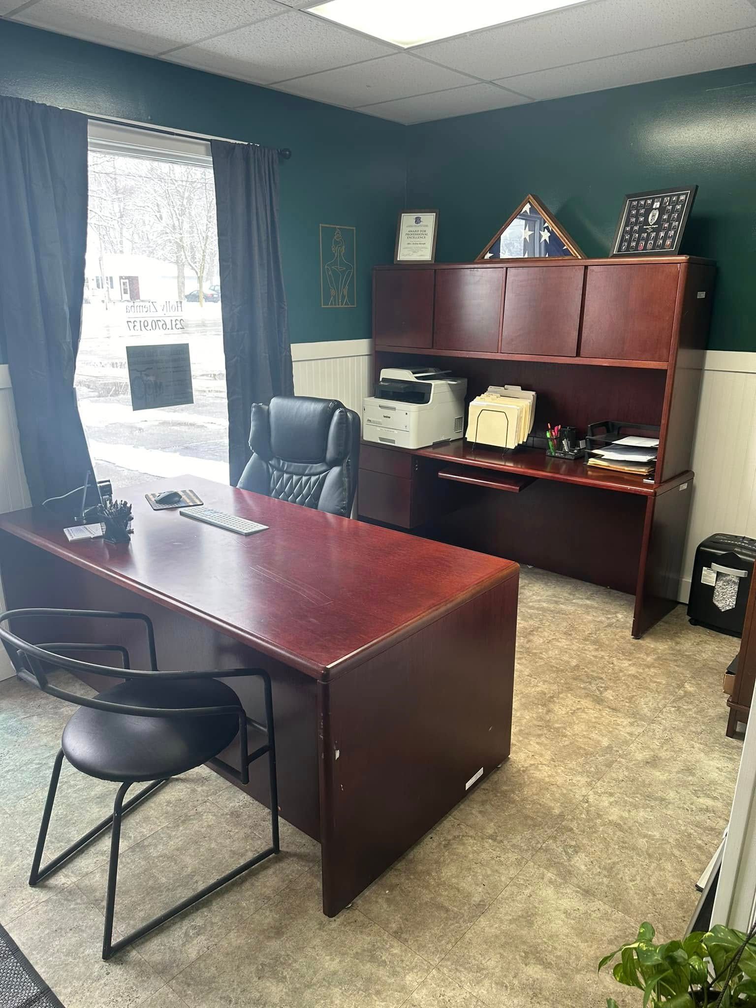 Small office with a wooden desk, black chair, shelving unit, and window with daylight.