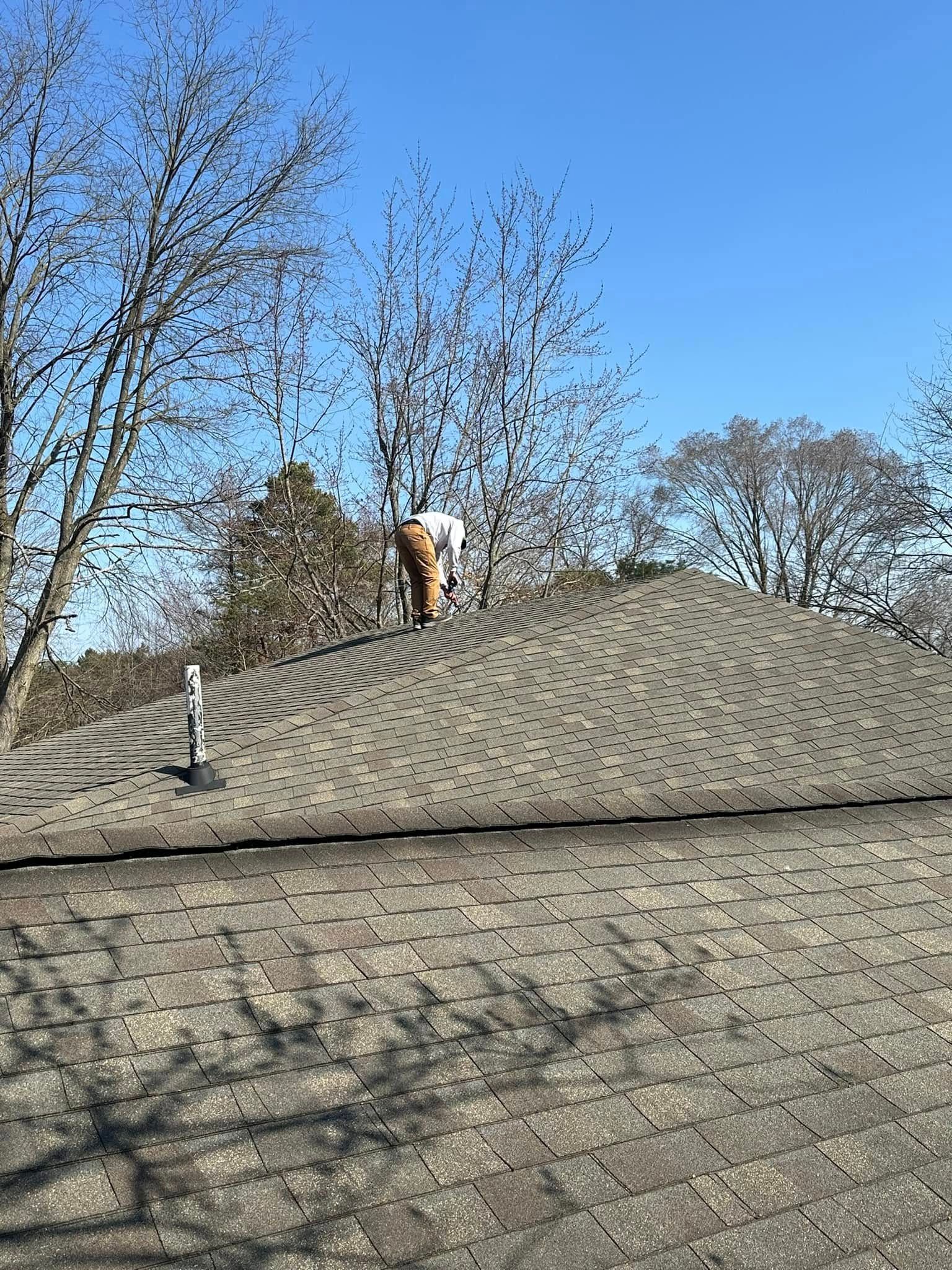 Person on a shingled roof beside leafless trees under a clear blue sky