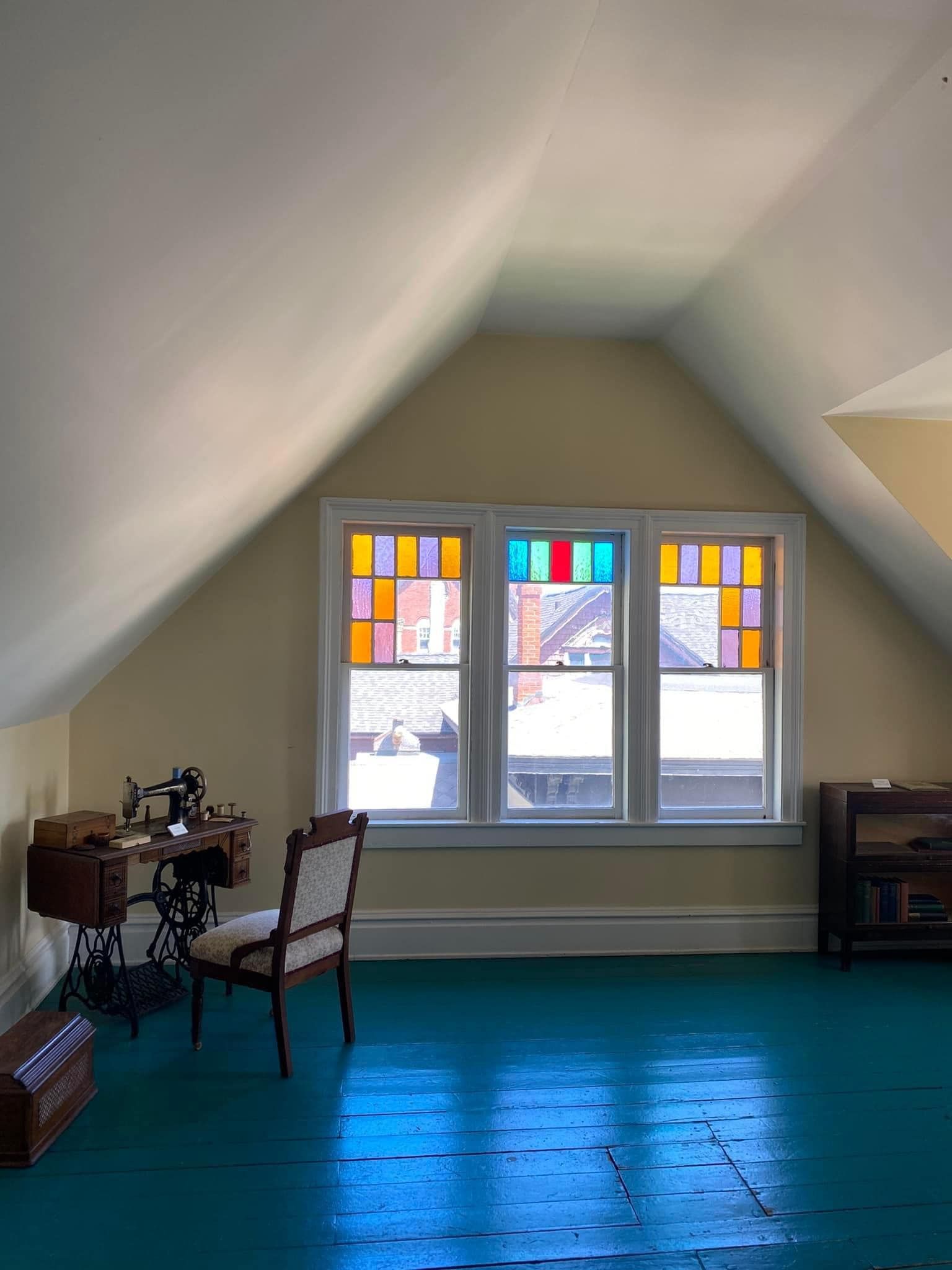 Attic room with sloped white ceiling, stained-glass windows, teal carpet, chair, and sewing machine table.