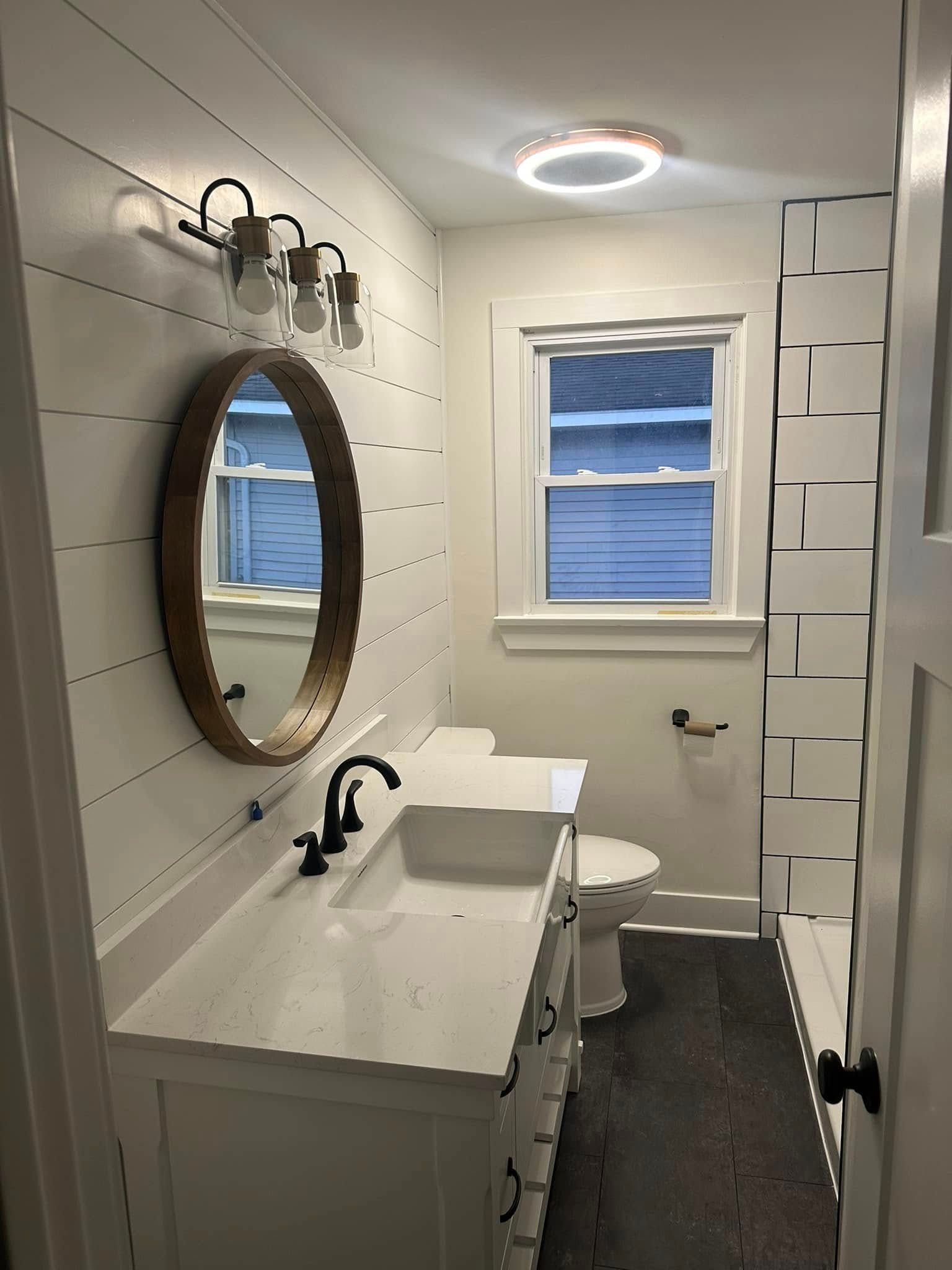 Small white bathroom with round mirror, sink, toilet, window, and ceiling light.