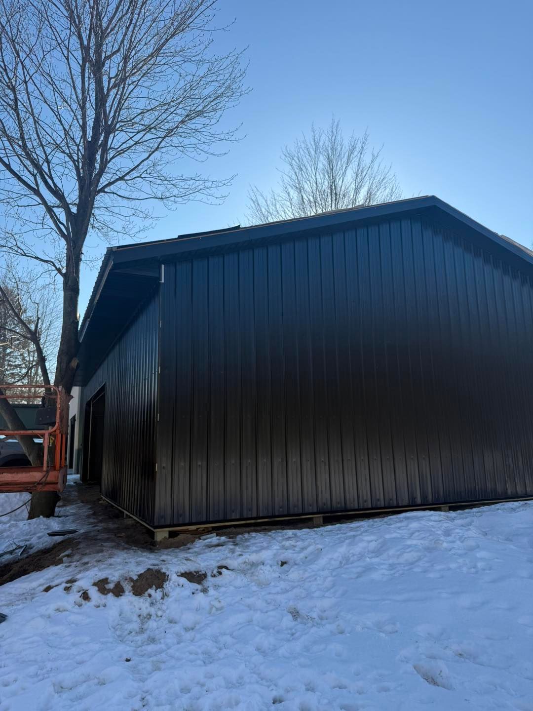 Dark shed with vertical siding beside snowy ground and leafless trees on a winter day
