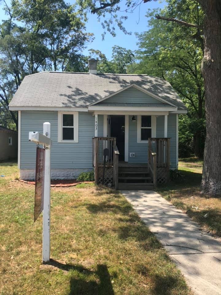 Small blue house with a front porch, steps, and a mailbox by a sidewalk in a sunny yard.