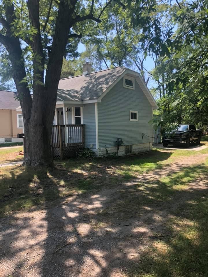 Small green house with a porch, large trees, and a parked car in a shaded yard