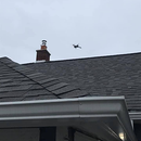 Small drone flying above a gray shingled roof and chimney under an overcast sky