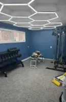 Small blue gym room with hexagon ceiling lights, dumbbells, a cable machine, and cleaning tools on the floor