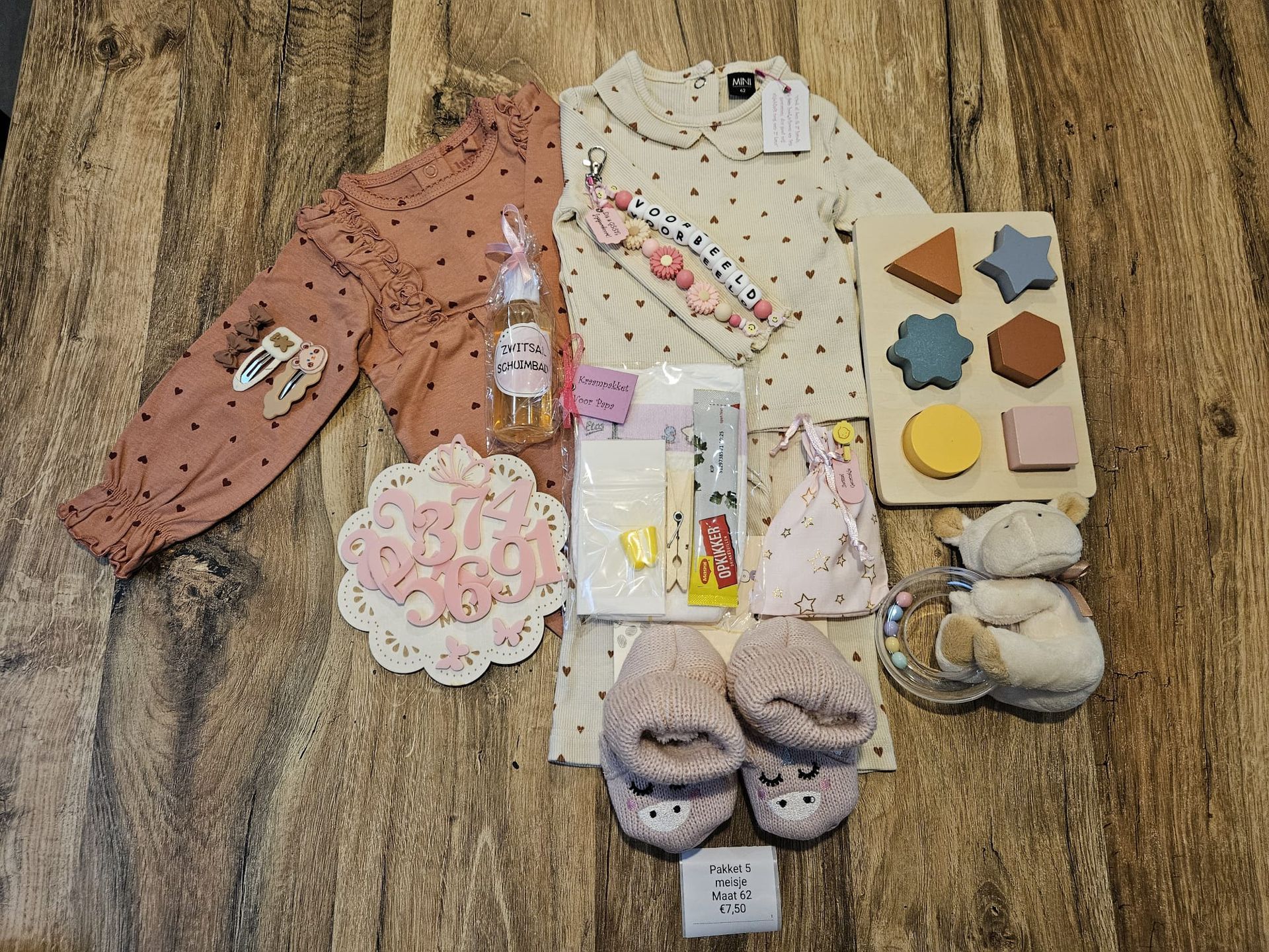 Een platte legging van babykleertjes, sokken, speelgoed en accessoires, uitgestald op een houten vloer.