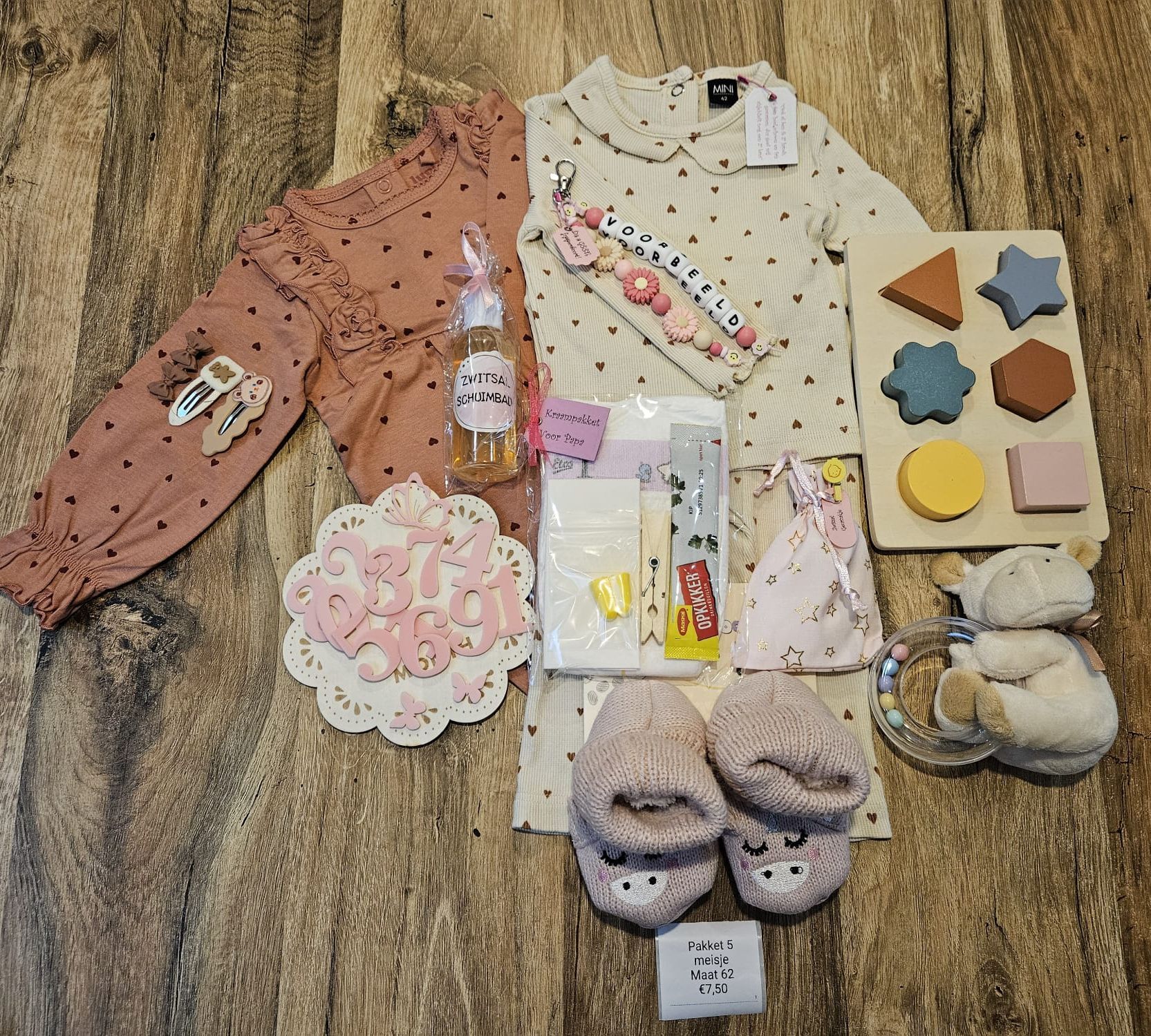 Kinderkleding en cadeautjes liggen uitgestald op een kleed, waaronder roze kleren, schoenen, speelgoed en een flesje.