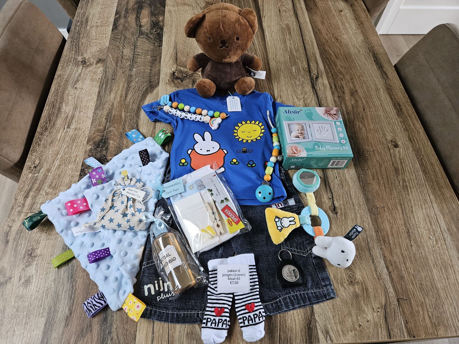 Babykleertjes, een knuffelbeer en opgevouwen dekens liggen uitgestald op een houten tafel.