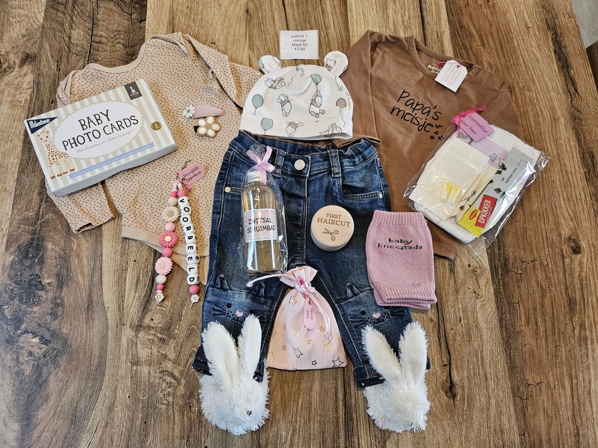 Platte legging van babykleertjes en -accessoires op een beige achtergrond, waaronder een konijnenpakje, schoentjes, een flesje en tasjes.