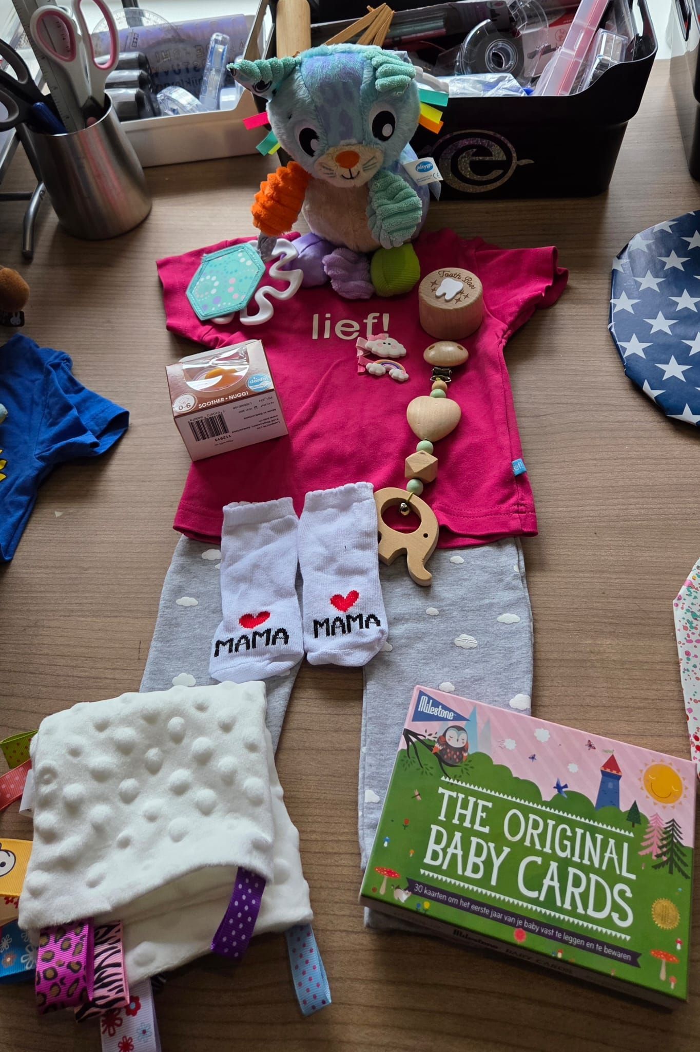 Een cadeauset voor een babypop, compleet met speelgoedkleertjes, fopspeen, flesje en babykaartjes, ligt op tafel.