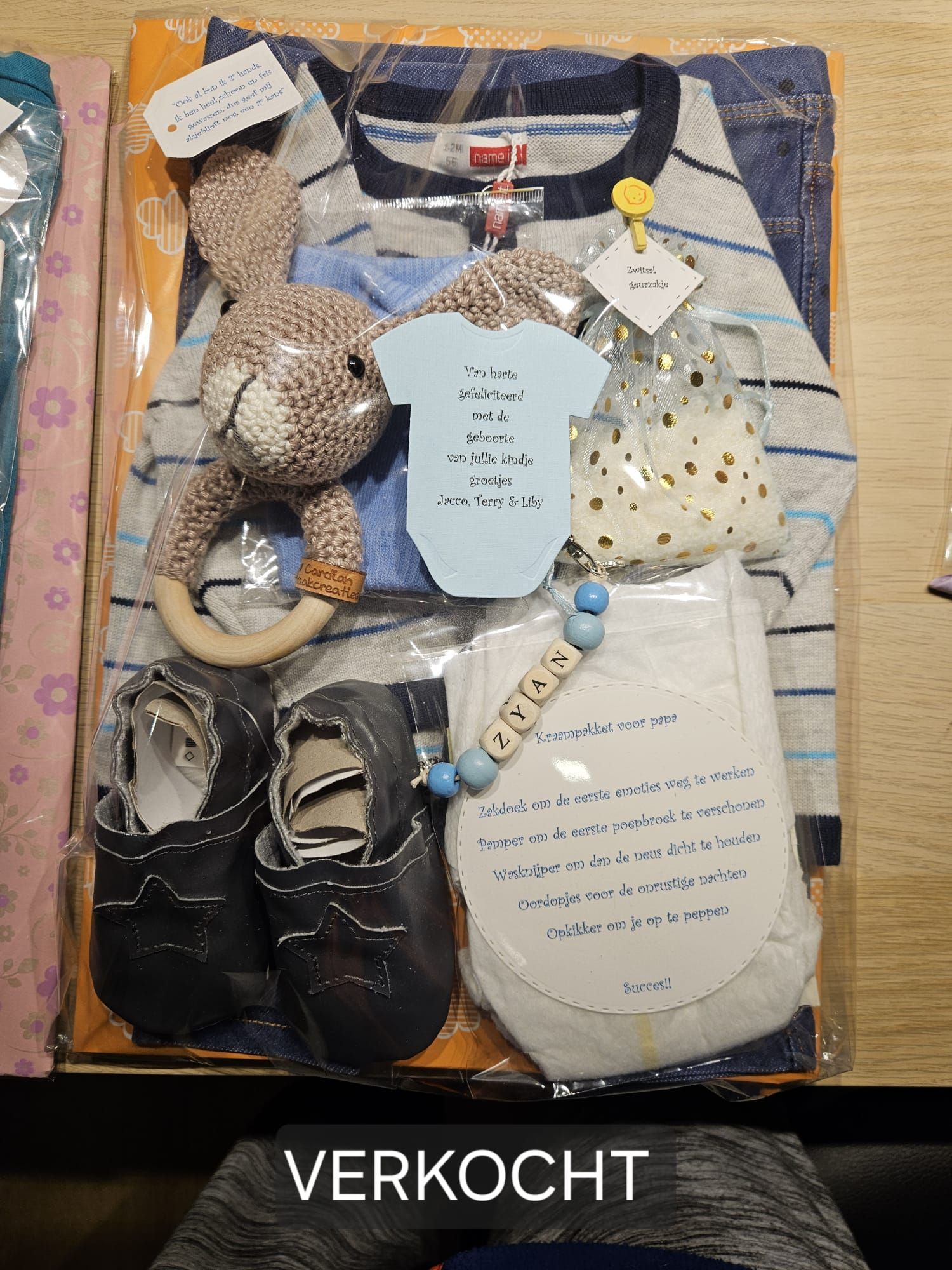 Diverse babykleertjes en -schoentjes in een geschenkmand op een gestreepte deken, onderaan gemerkt met 
