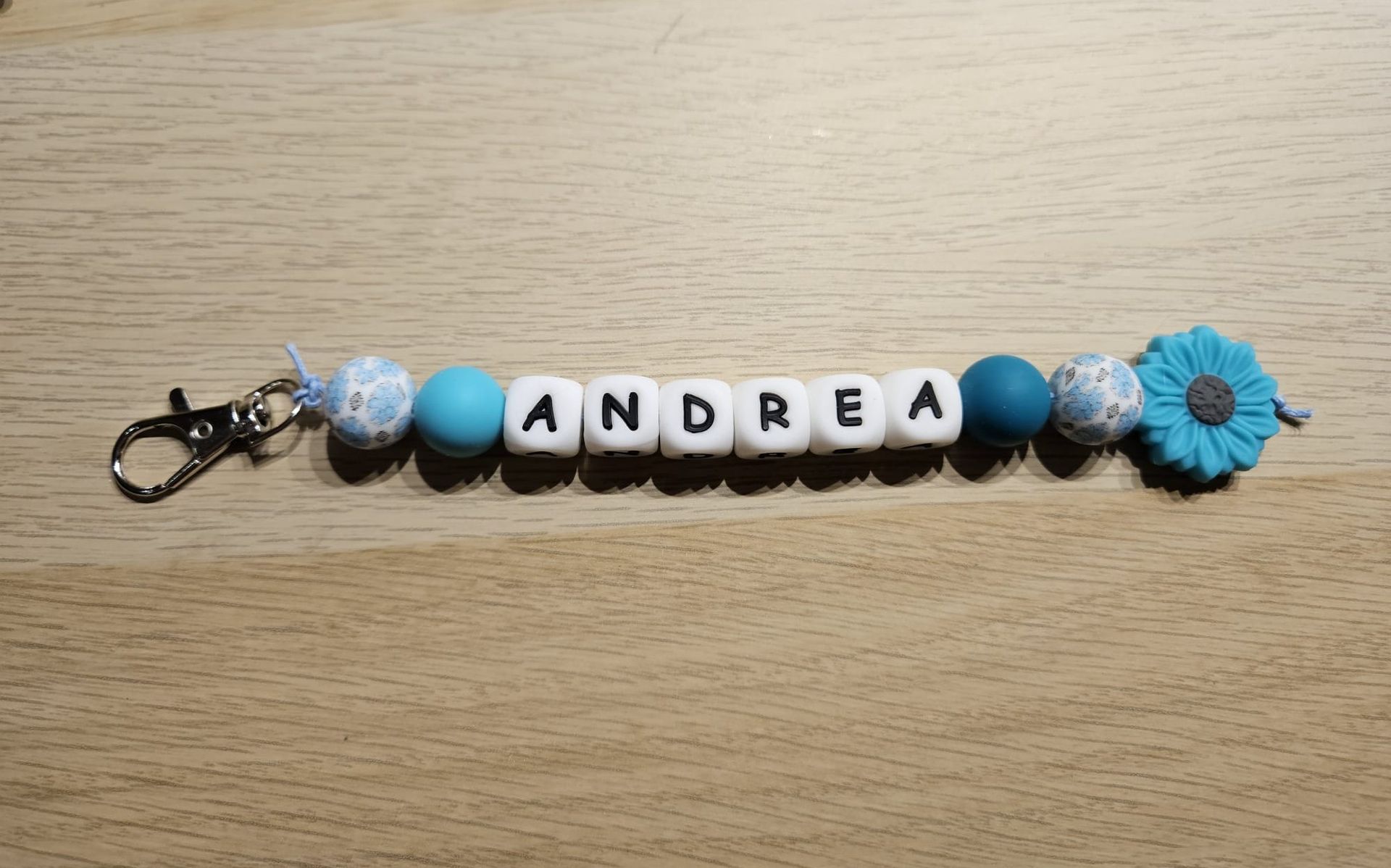 Blauw-witte kralen sleutelhanger met de naam ANDREA en een bloemhangertje op een houten ondergrond.