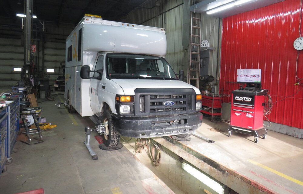 Camping-car blanc sur un pont élévateur dans un garage, en cours de réglage de la géométrie. Parois et équipement rouges visibles.